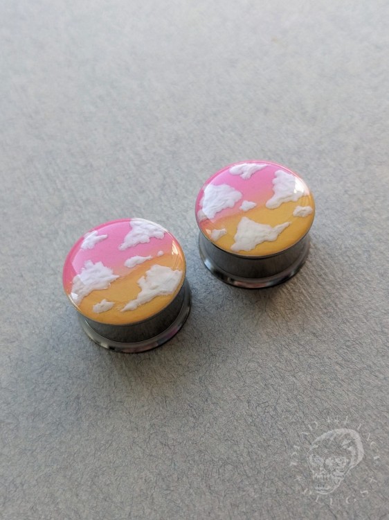 Silver ear plugs with pink and yellow ombre effect, decorated with fluffy white clouds.